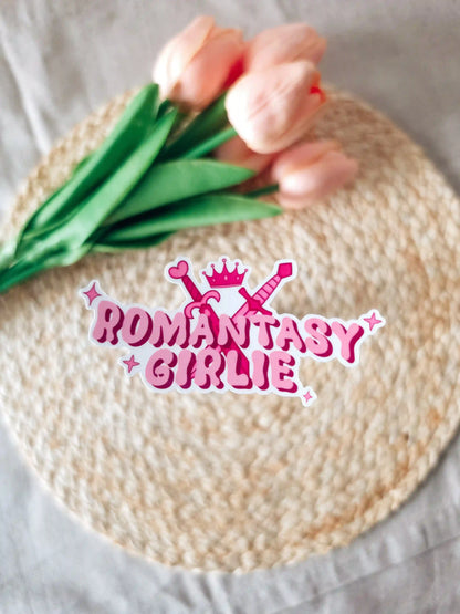 A die-cut sticker reading "Romantasy Girlie" in bold pink bubbly lettering, decorated with two crossed swords, a pink crown, a heart, and sparkle details, placed on a woven rattan mat with peach roses and green leaves resting in the upper corner.