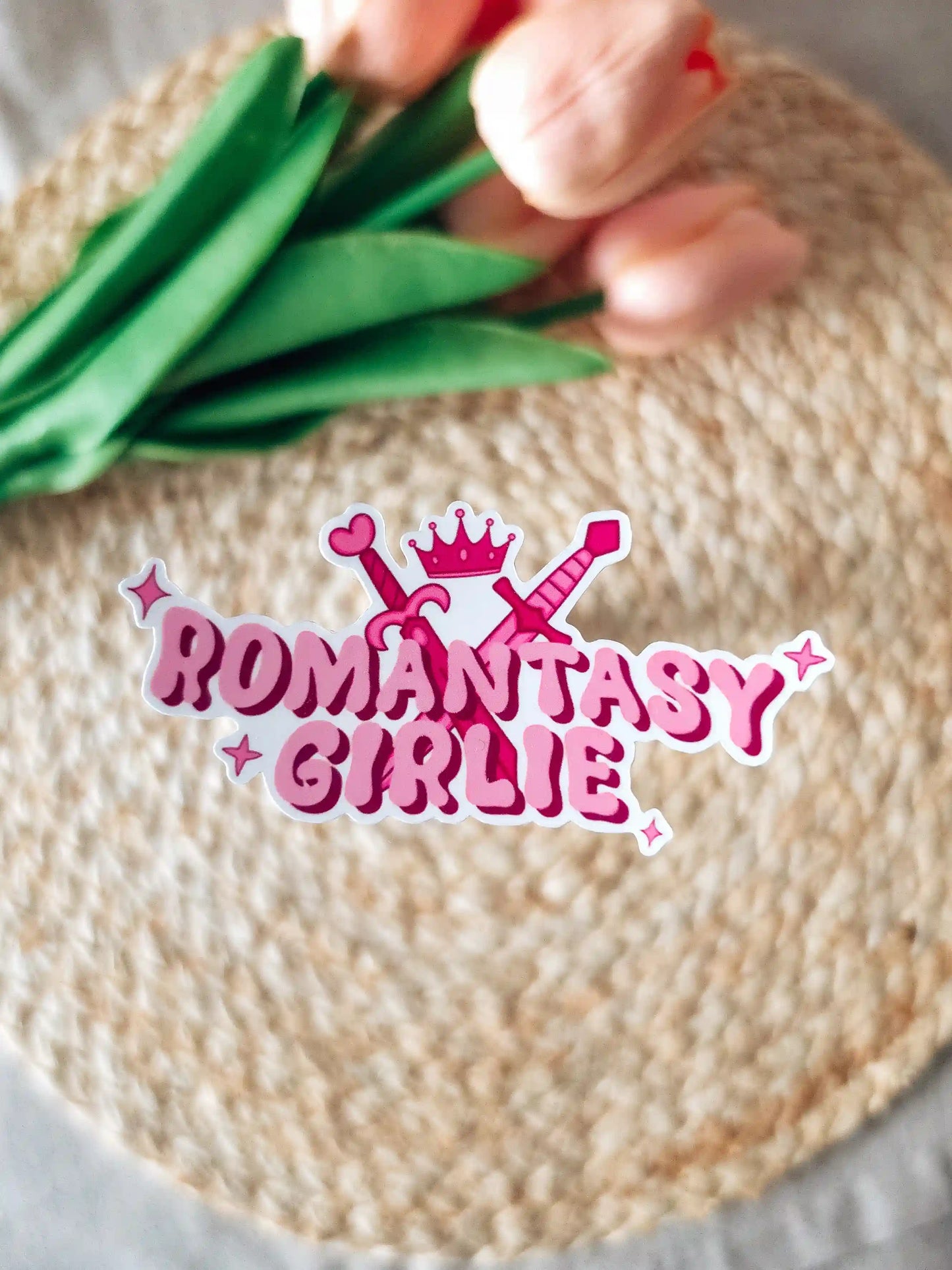 A die-cut sticker reading "Romantasy Girlie" in bold pink bubbly lettering, decorated with two crossed swords, a pink crown, a heart, and sparkle details, placed on a woven rattan mat with peach roses and green leaves resting in the upper corner.
