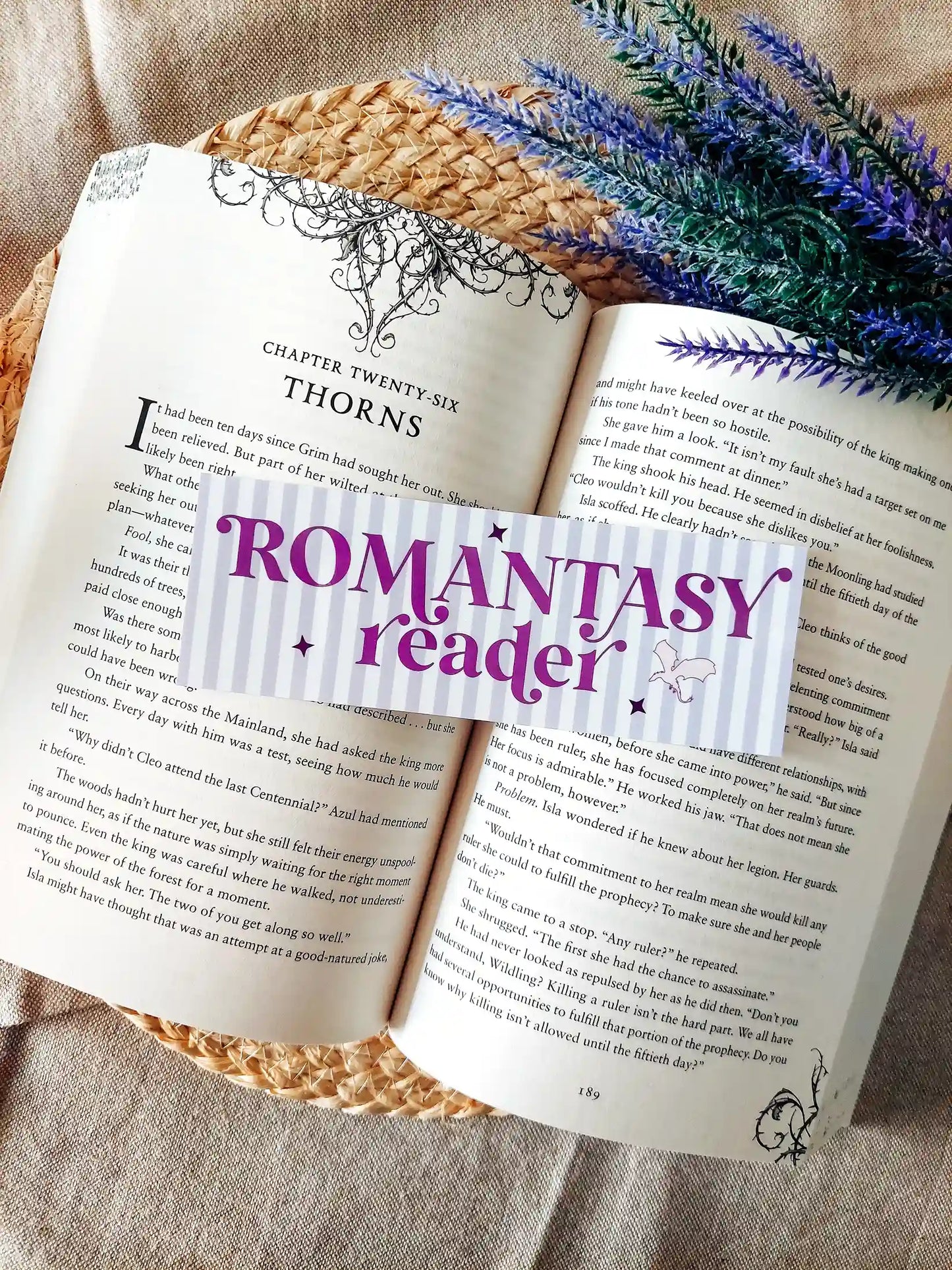 Illustrated bookmark with the text "Romantasy Reader", featuring purple stripes and decorative stars and dragons, resting on an open book.