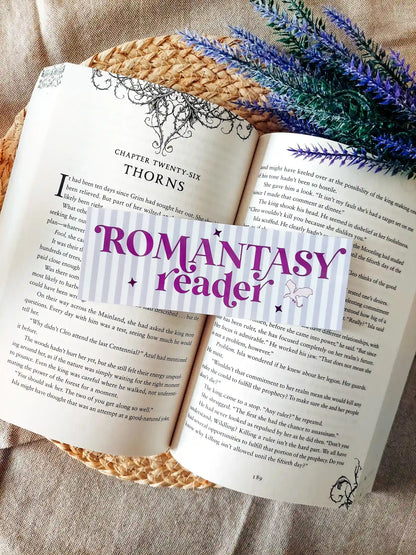 Illustrated bookmark with the text "Romantasy Reader", featuring purple stripes and decorative stars and dragons, resting on an open book.