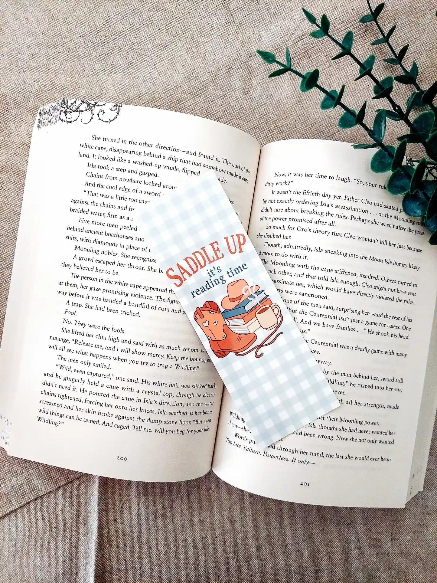 Bookmark with the text "Saddle Up, it’s reading time", featuring a western-themed design with cowboy boots, a hat, stacked books and a mug on a soft gingham background, resting on an open book.