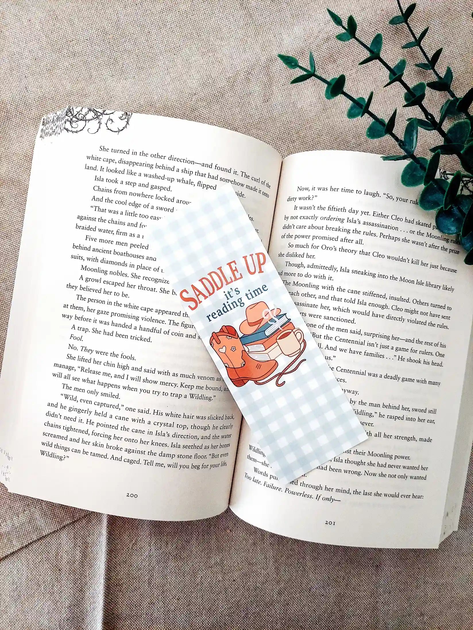 Bookmark with the text "Saddle Up, it’s reading time", featuring a western-themed design with cowboy boots, a hat, stacked books and a mug on a soft gingham background, resting on an open book.