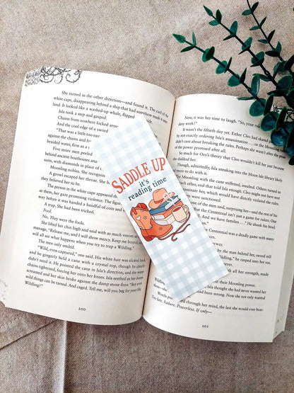 Bookmark with the text "Saddle Up, it’s reading time", featuring a western-themed design with cowboy boots, a hat, stacked books and a mug on a soft gingham background, resting on an open book.