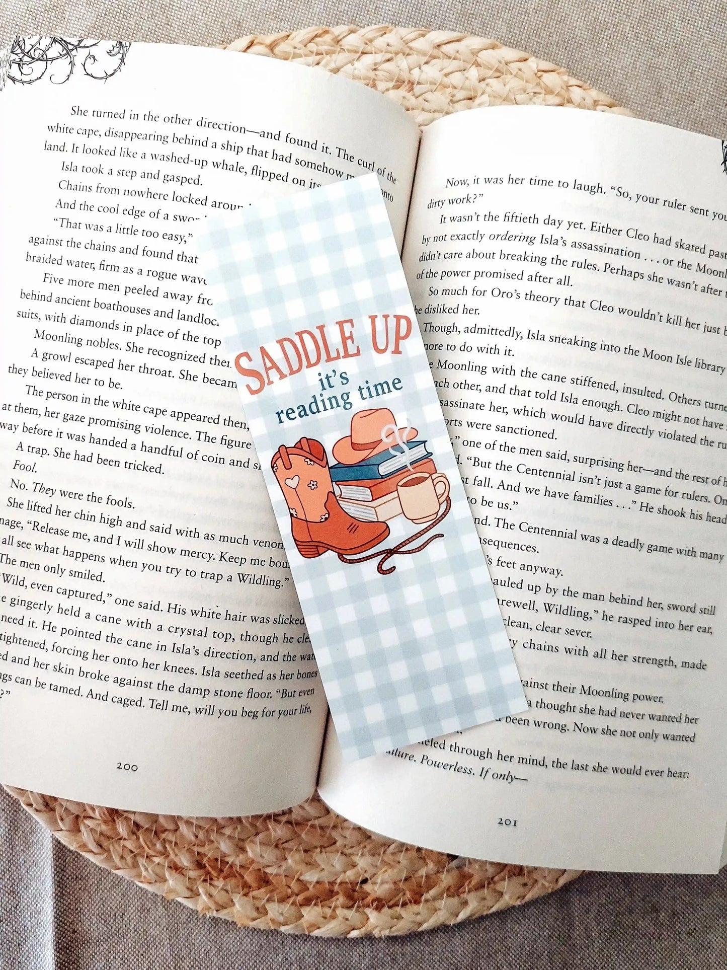 Bookmark with the text "Saddle Up, it’s reading time", featuring a western-themed design with cowboy boots, a hat, stacked books and a mug on a soft gingham background, resting on an open book.