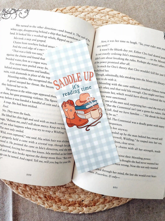 Bookmark with the text "Saddle Up, it’s reading time", featuring a western-themed design with cowboy boots, a hat, stacked books and a mug on a soft gingham background, resting on an open book.