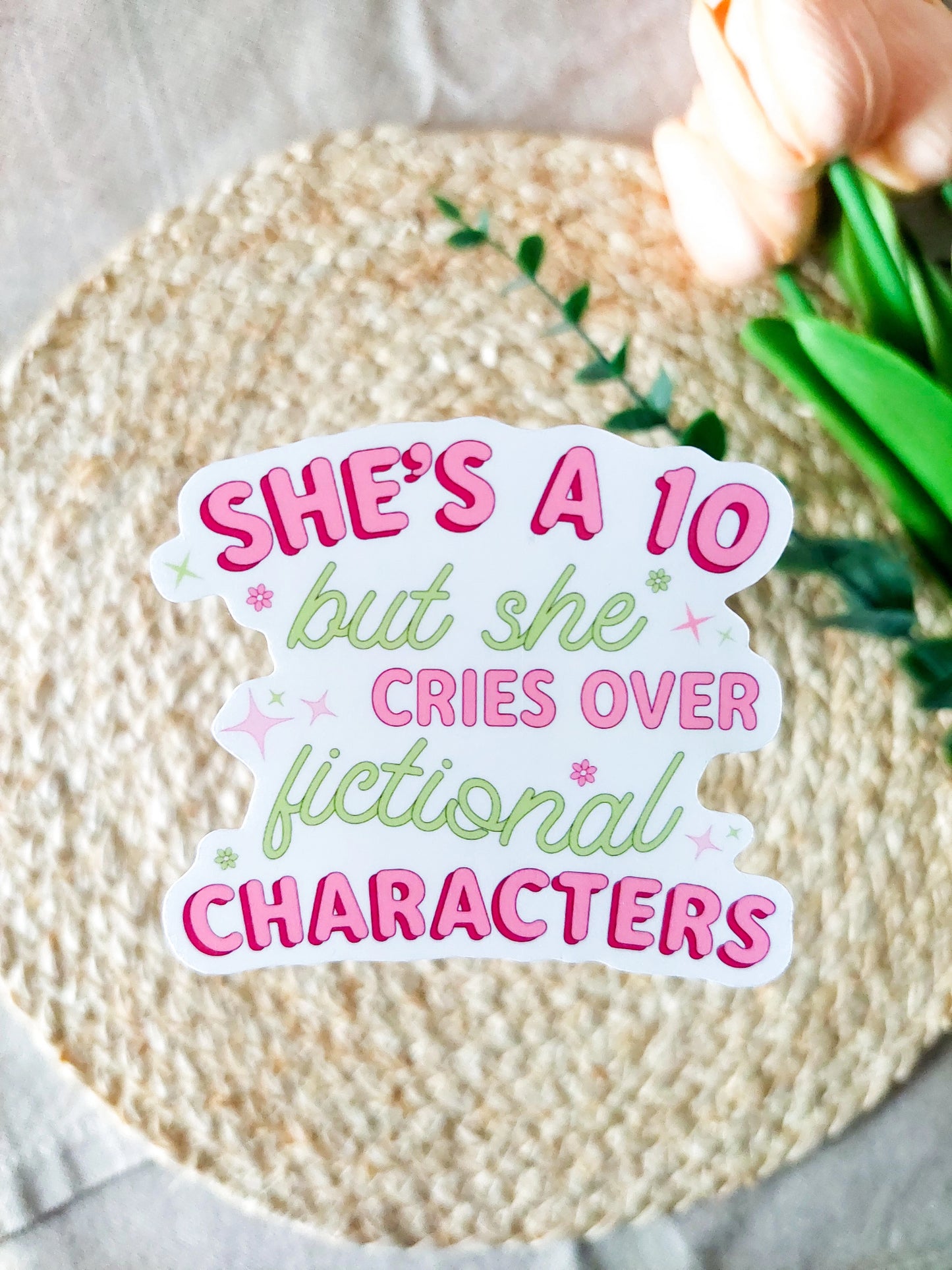 "She's A 10 But She Cries Over Fictional Characters" sticker on a rattan placemat, featuring pink and green text with small flowers and stars detail. 