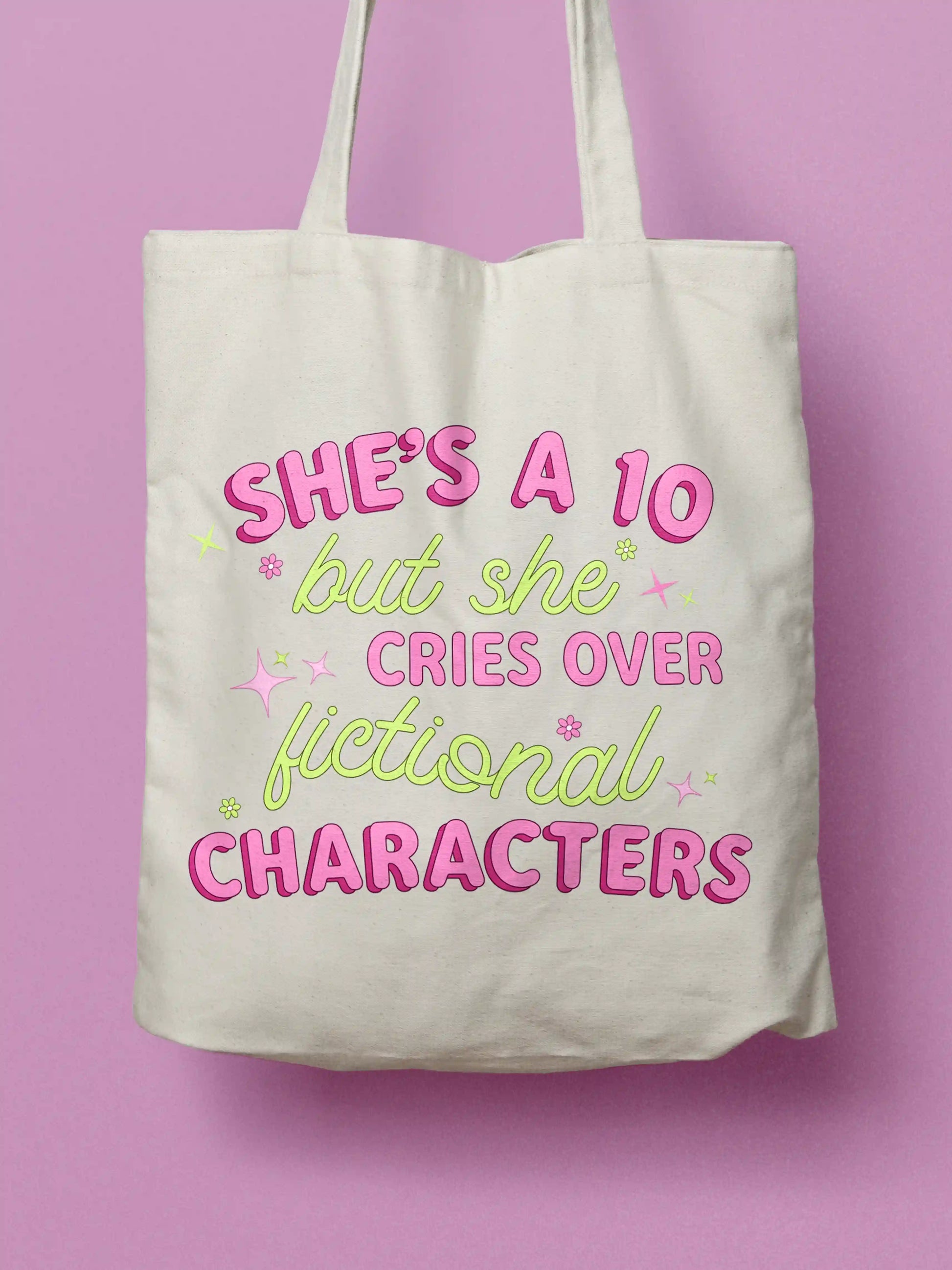 An white canvas tote bag with pink and lime green text that reads "She's A 10 But She Cries Over Fictional Characters"' with star and flower details, on a pink background.

