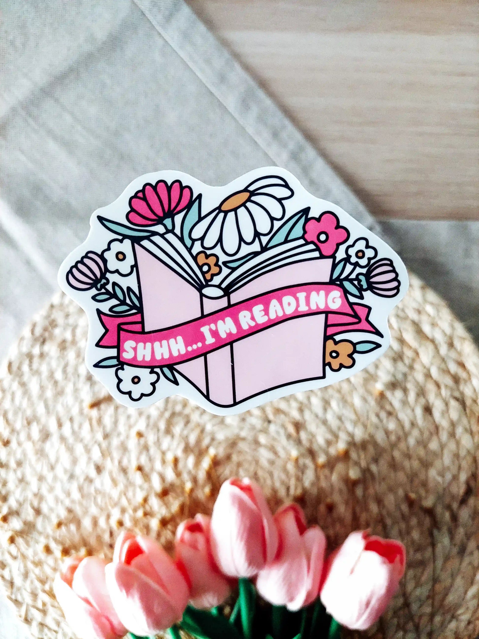 Vinyl sticker featuring an open book with flowers and a pink banner with the text "Shhh I’m Reading", photographed on neutral fabric background with woven mat and pink tulips.