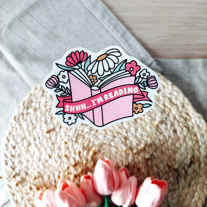 Vinyl sticker featuring an open book with flowers and a pink banner with the text "Shhh I’m Reading", photographed on neutral fabric background with woven mat and pink tulips.
