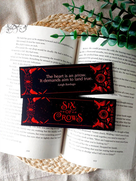Bookmark inspired by the Six of Crows collector edition, featuring the quote "The heart is an arrow. It demands aim to land true." in red on a black background, resting on an open book.