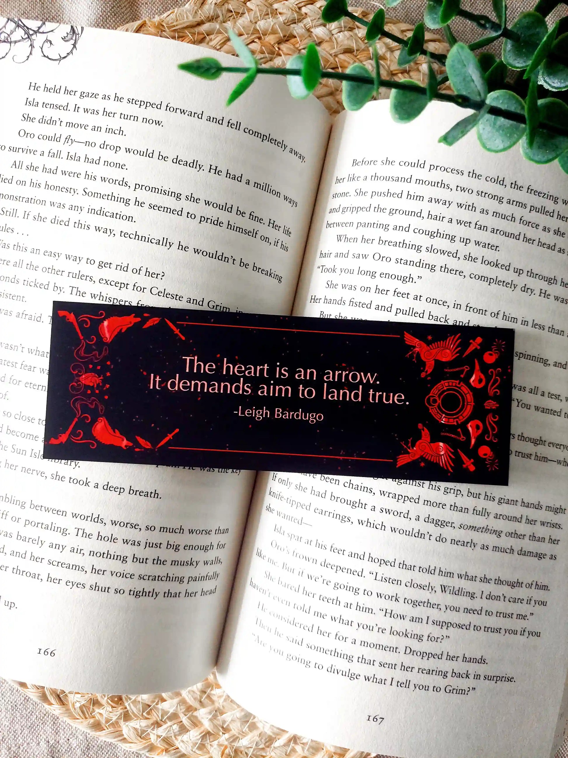 Bookmark inspired by the Six of Crows collector edition, featuring the quote "The heart is an arrow. It demands aim to land true." in red on a black background, resting on an open book.
