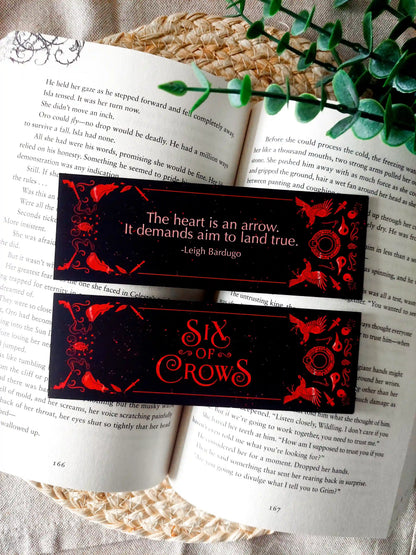Bookmark inspired by the Six of Crows collector edition, featuring the quote "The heart is an arrow. It demands aim to land true." in red on a black background, resting on an open book.