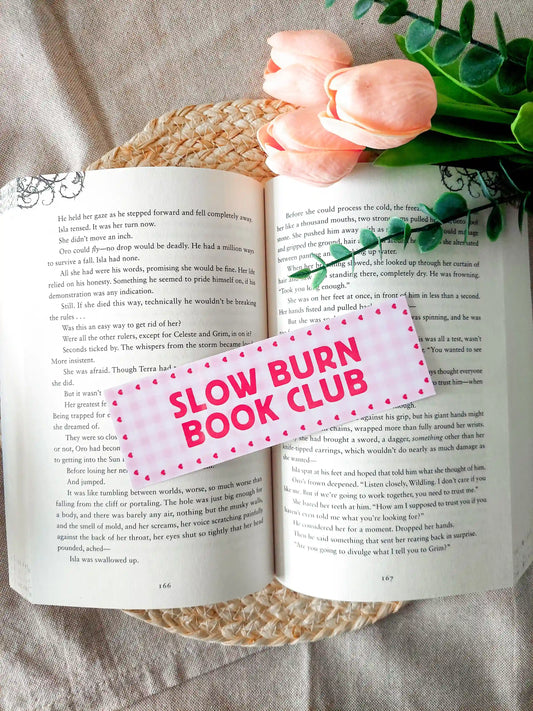 Bookmark with the text "Slow Burn Book Club", featuring a pink gingham pattern with small heart details, resting on an open book. 