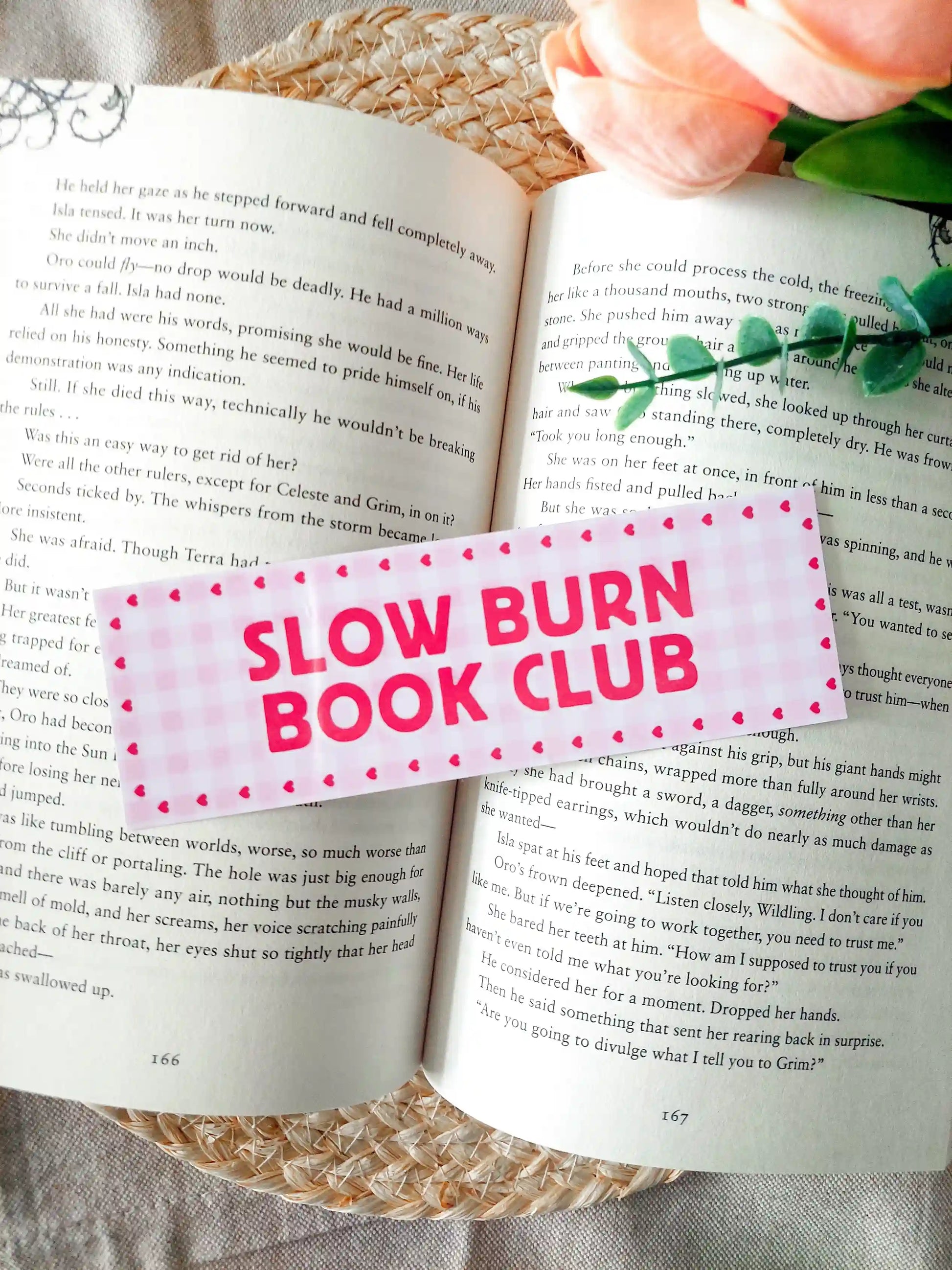 Bookmark with the text "Slow Burn Book Club", featuring a pink gingham pattern with small heart details, resting on an open book. 