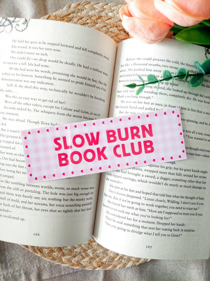 Bookmark with the text "Slow Burn Book Club", featuring a pink gingham pattern with small heart details, resting on an open book. 