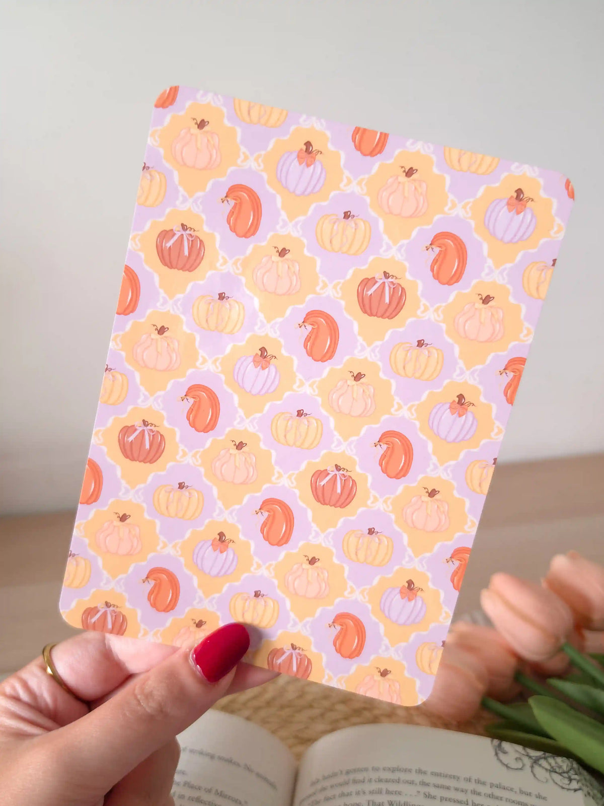 A laminated e-reader insert featuring a tile pattern with illustrated pumpkins in pastel lavender, peach, and orange tones, resting on an open book on a woven rattan mat, with green leafy branches visible in the lower left corner.