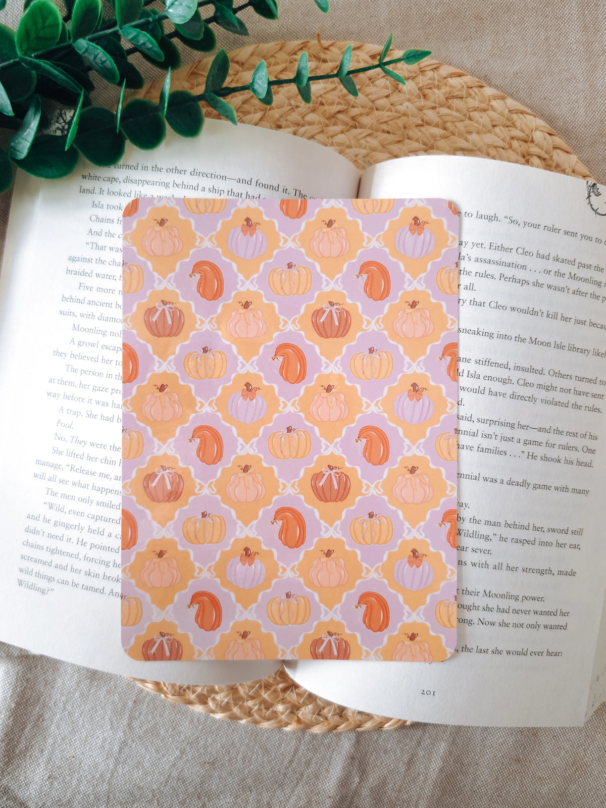 A laminated e-reader insert featuring a tile pattern with illustrated pumpkins in pastel lavender, peach, and orange tones, resting on an open book on a woven rattan mat, with green leafy branches visible in the lower left corner.