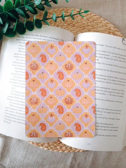 A laminated e-reader insert featuring a tile pattern with illustrated pumpkins in pastel lavender, peach, and orange tones, resting on an open book on a woven rattan mat, with green leafy branches visible in the lower left corner.