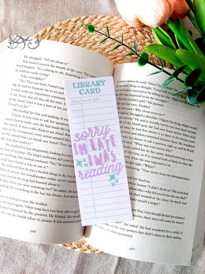 Bookmark designed like a library card with pastel colors and the text "Sorry I'm late, I was reading", placed on an open book.