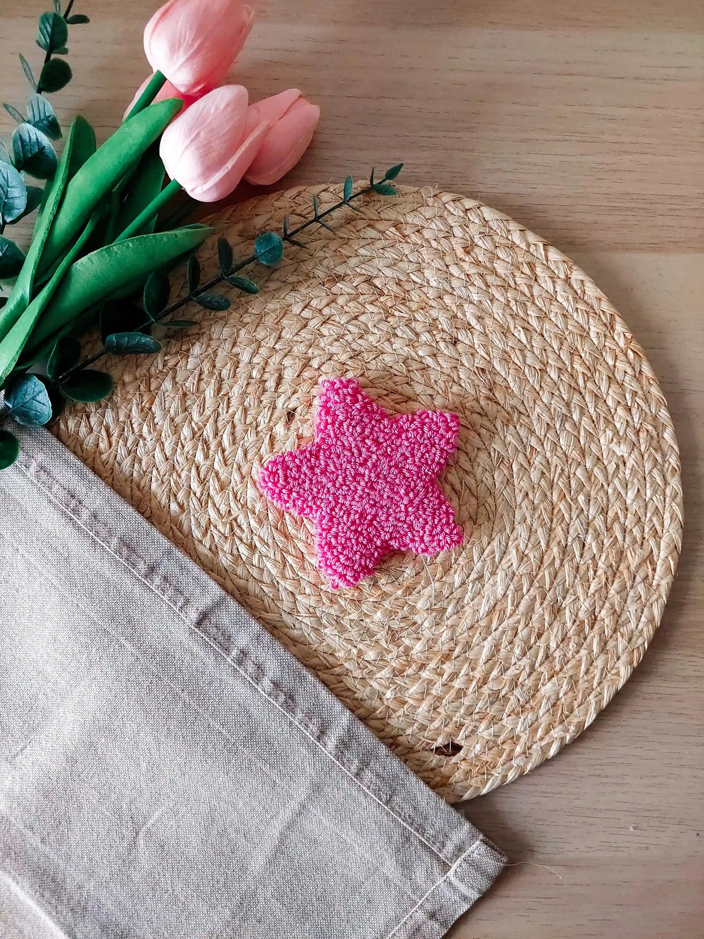 Hot pink star shaped punch needle coaster photographed on a woven mat with decorative pink flowers in the back.