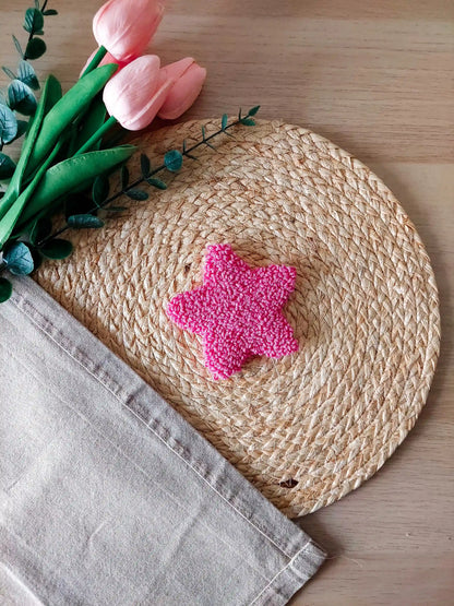 Hot pink star shaped punch needle coaster photographed on a woven mat with decorative pink flowers in the back.