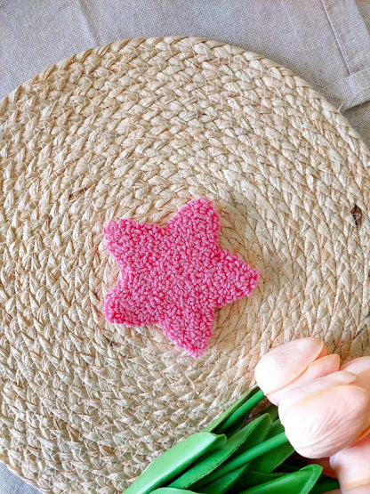 Hot pink star shaped punch needle coaster photographed on a woven mat with decorative pink flowers in the back.