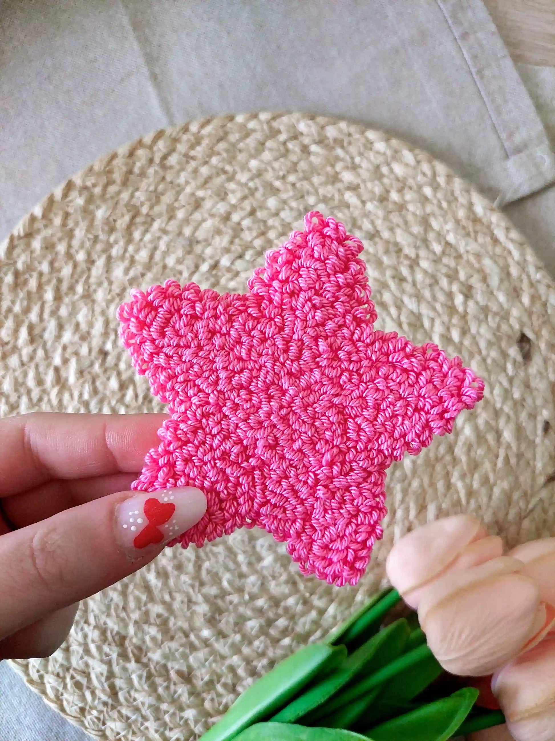 Hot pink star shaped punch needle coaster photographed on a woven mat with decorative pink flowers in the back.
