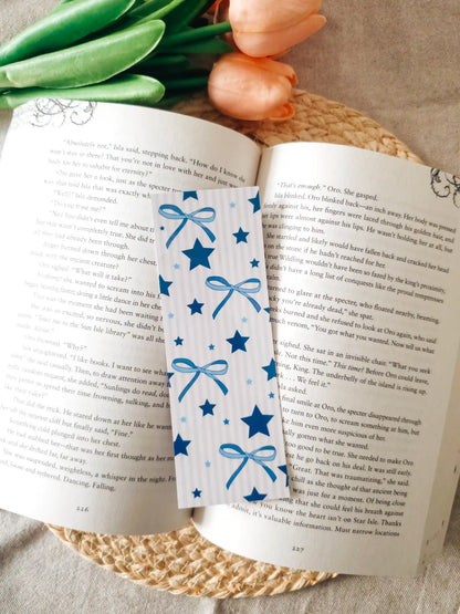 An open book spread on a woven mat with green foliage around it. A decorative bookmark with blue ribbon bows and stars is placed across the pages.