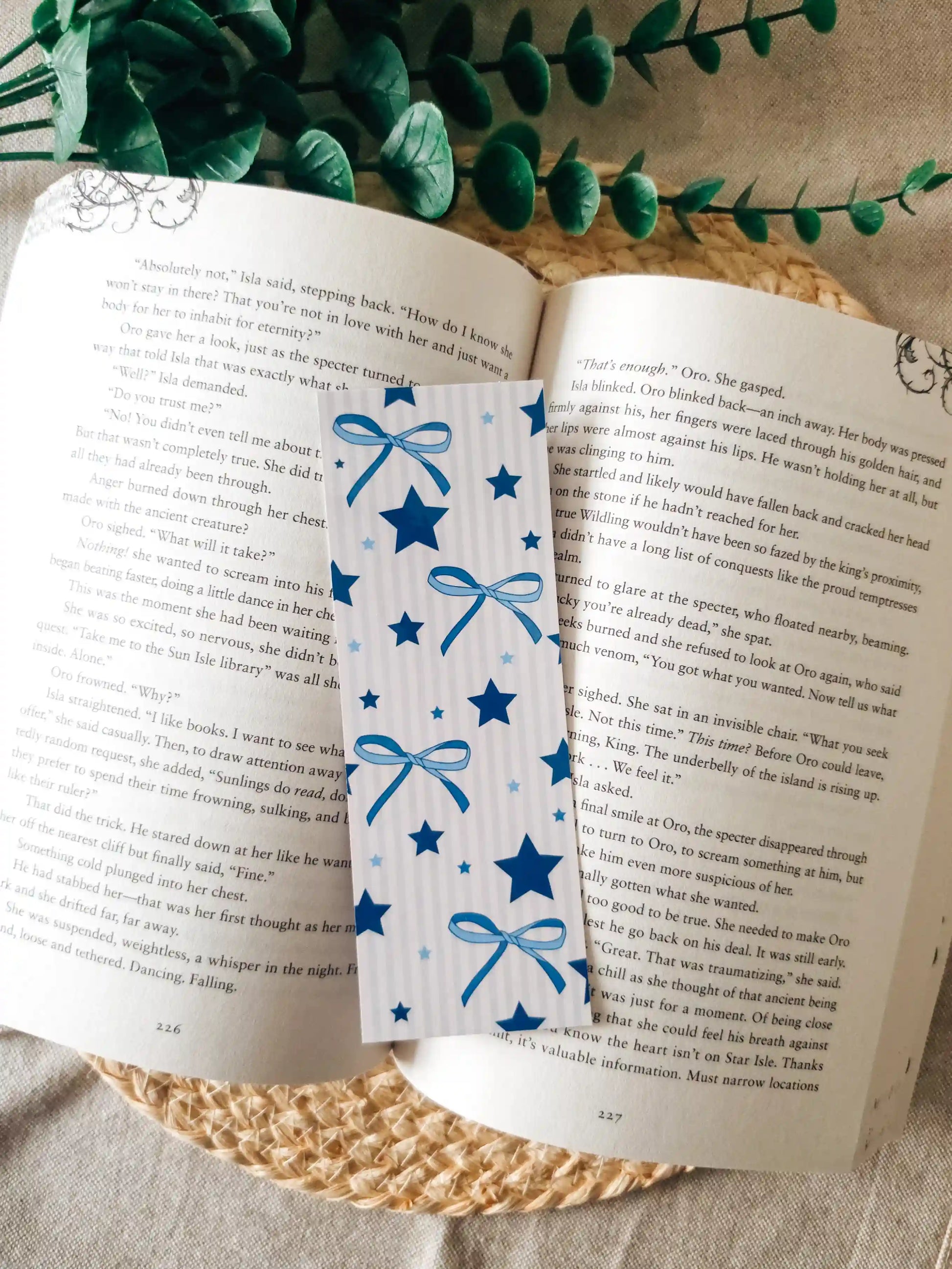 An open book spread on a woven mat with green foliage around it. A decorative bookmark with blue ribbon bows and stars is placed across the pages.