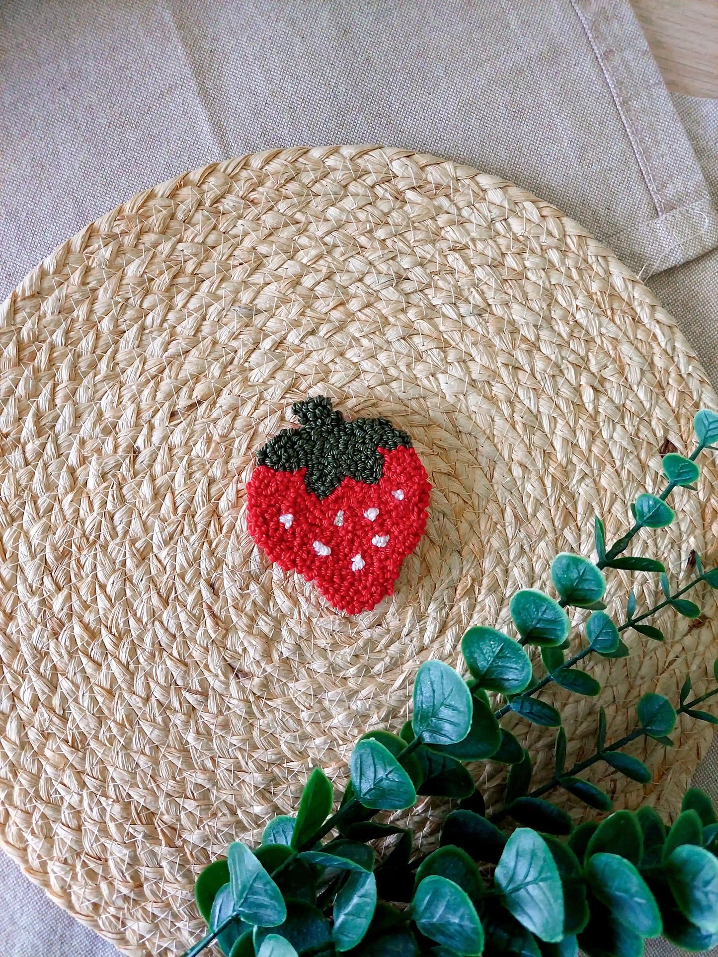 Handmade strawberry-shaped punch needle coaster in red and dark green yarn, photographed on a woven mat with decorative flowers in the back.