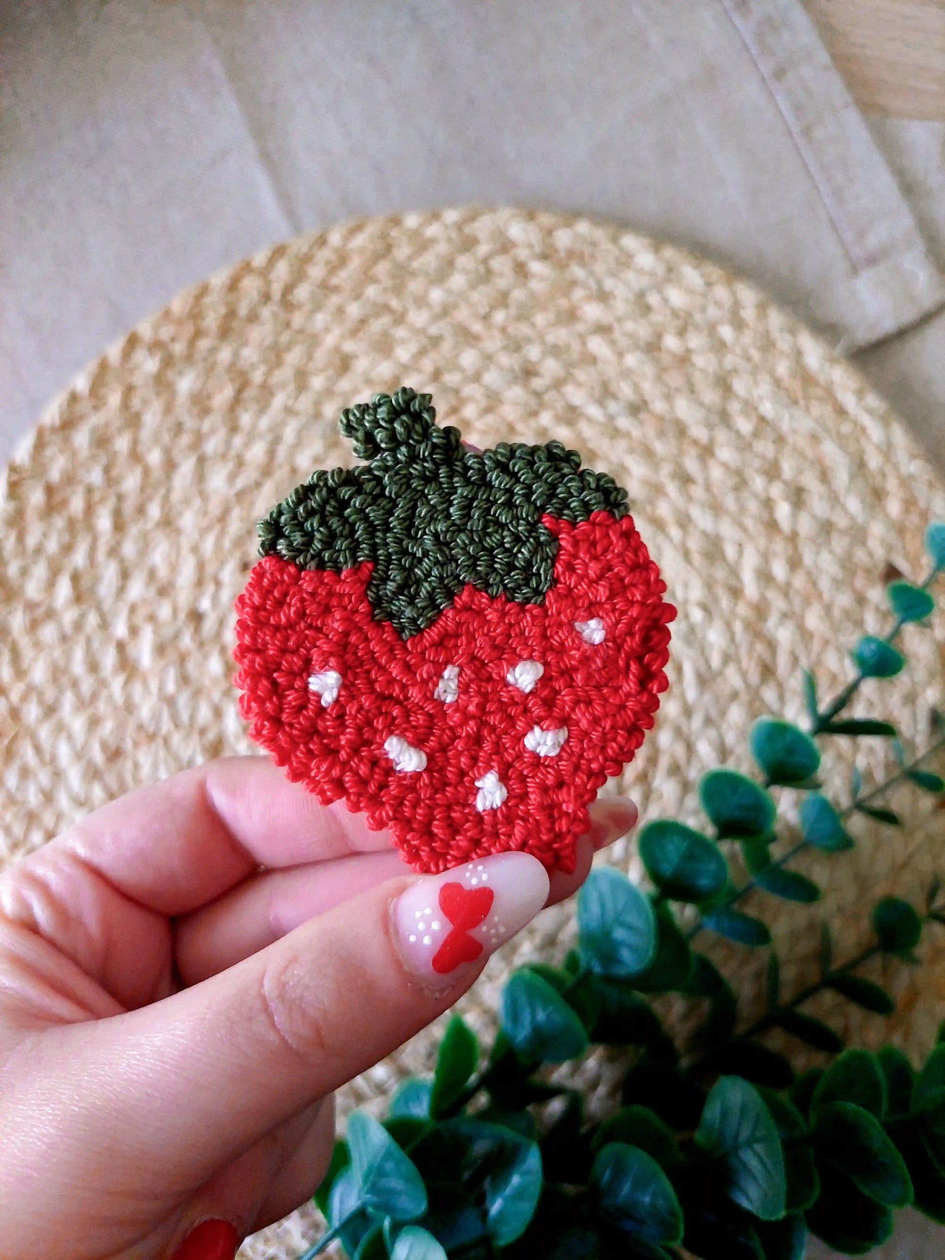 Handmade strawberry-shaped punch needle coaster in red and dark green yarn, photographed on a woven mat with decorative flowers in the back.