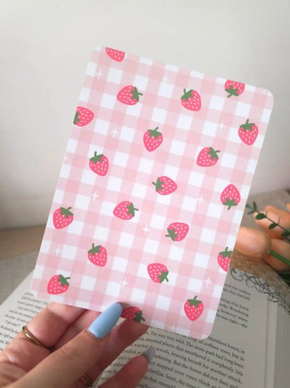 A hand holding an e-reader insert with a soft pink gingham background featuring pink strawberry illustrations and small sparkle details scattered across it.