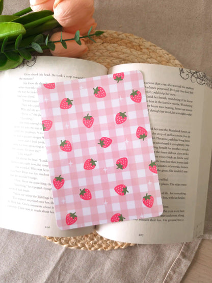 An e-reader insert with a soft pink gingham background featuring pink strawberry illustrations and small sparkle details scattered across it, placed on an open book resting on a woven rattan mat. 