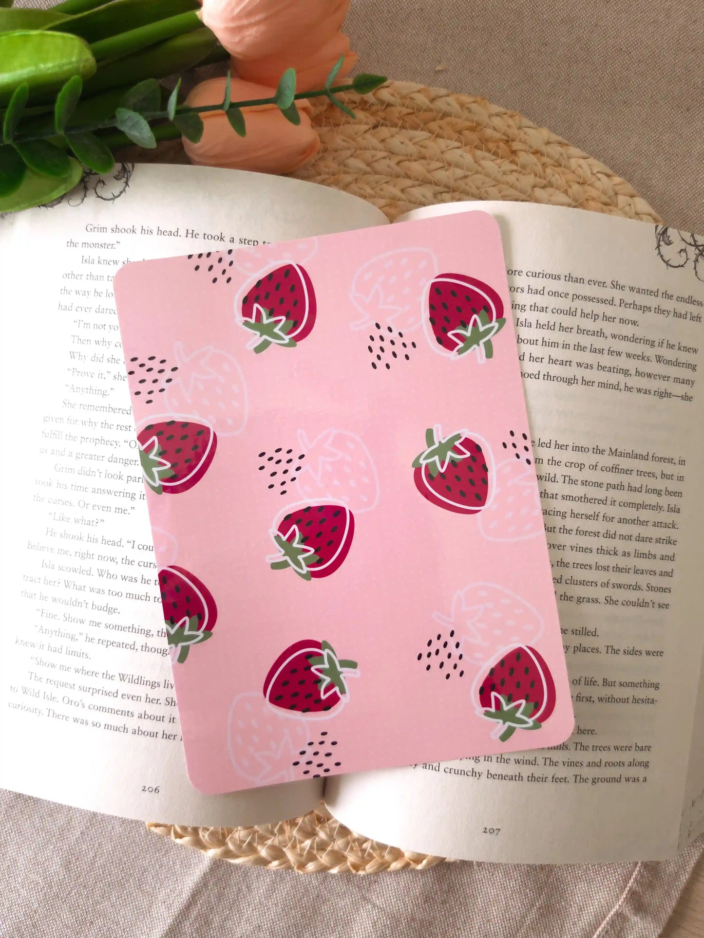 An e-reader insert with a solid pink background featuring red strawberry illustrations with white outlines and green leaves, with small black dot details scattered across it, placed on an open book resting on a woven rattan mat.
