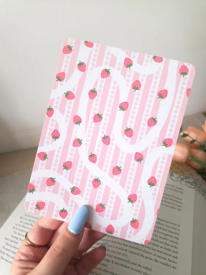 A hand holding an e-reader insert with a pink striped background featuring a wavy white ribbon design overlay and small pink strawberry illustrations scattered across it.