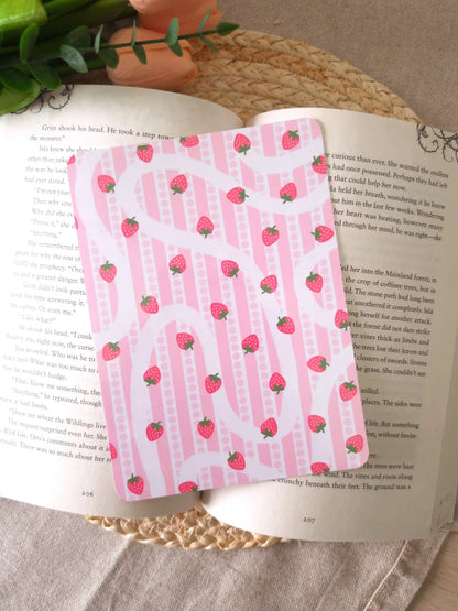 An e-reader insert with a pink striped background featuring a wavy white ribbon design overlay and small pink strawberry illustrations scattered across it, placed on an open book resting on a woven rattan mat. 