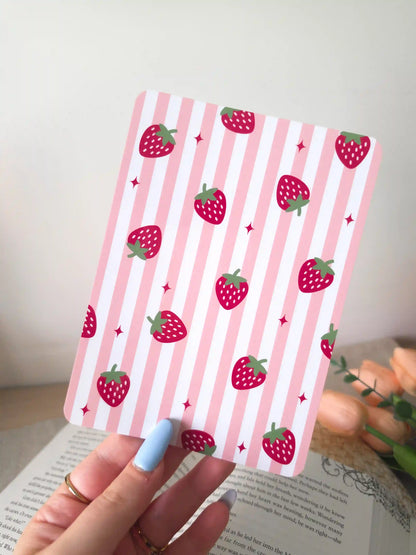 A hand holding an e-reader insert with a pink and white striped background featuring large red strawberry illustrations and small pink sparkle details scattered across it.