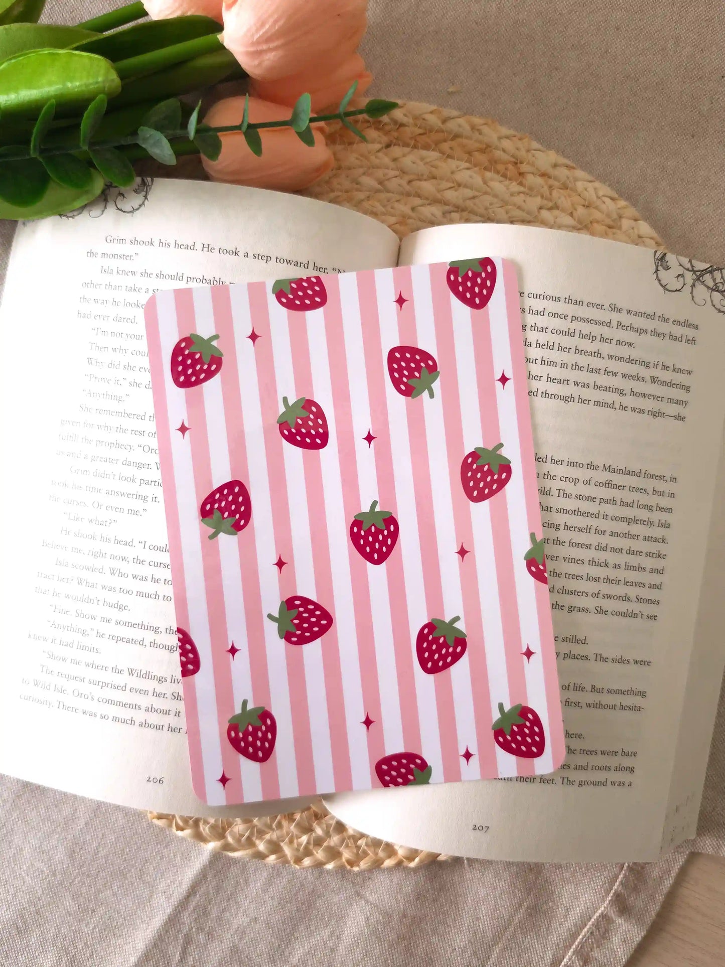An e-reader insert with a pink and white striped background featuring large red strawberry illustrations and small pink sparkle details scattered across it, placed on an open book resting on a woven rattan mat.