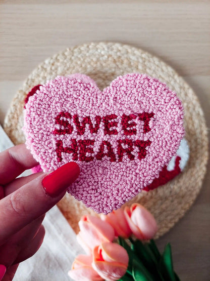 Handmade pink heart punch needle coaster with red "Sweetheart" lettering, displayed on a woven mat with decorative flowers in the back. 
