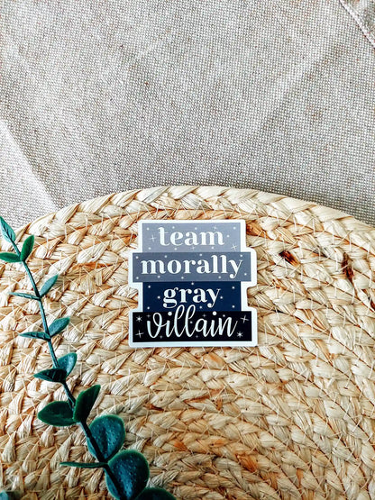 Vinyl sticker with the phrase "Team Morally Gray Villain" in layered black and gray text with small star details, photographed on a neutral textured background.