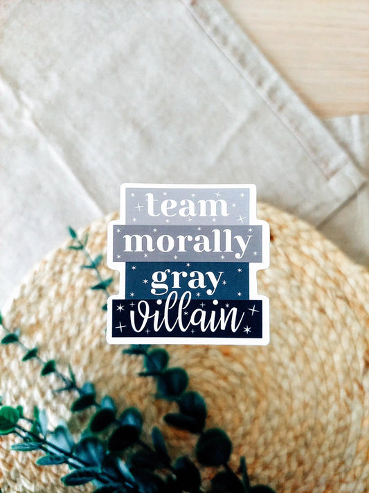 Vinyl sticker with the phrase "Team Morally Gray Villain" in layered black and gray text with small star details, photographed on a neutral textured background.