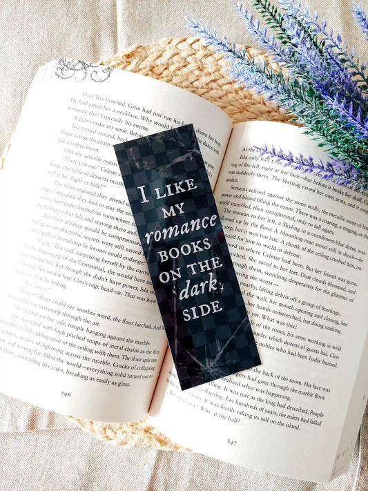 Book with a bookmark reading "I Like My Romance Books On The Dark Side" placed on an open book, featuring a dark checkered marble pattern with white text and brokeen glass details.