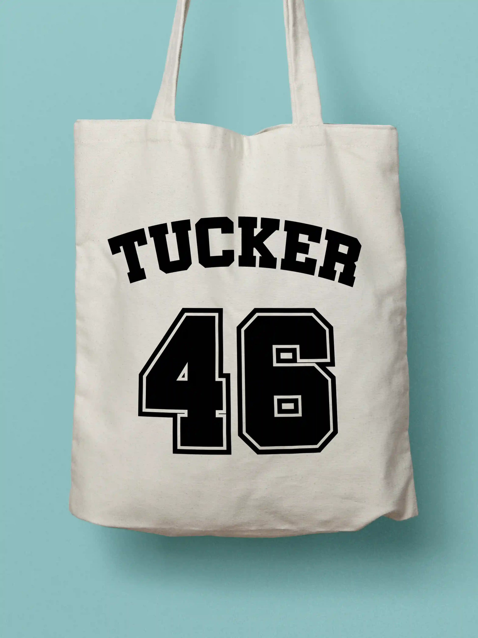 A white canvas tote bag featuring "Tucker 46" in bold black collegiate-style lettering, photographed against a lavender background.