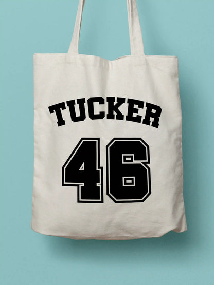 A white canvas tote bag featuring "Tucker 46" in bold black collegiate-style lettering, photographed against a lavender background.