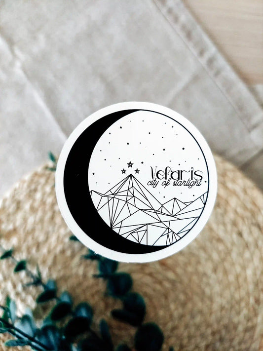 Round vinyl sticker featuring the phrase "Velaris city of starlight" with a crescent moon, stars and geometric mountain illustration inspired by ACOTAR's Velariss, photographed on a woven background.

