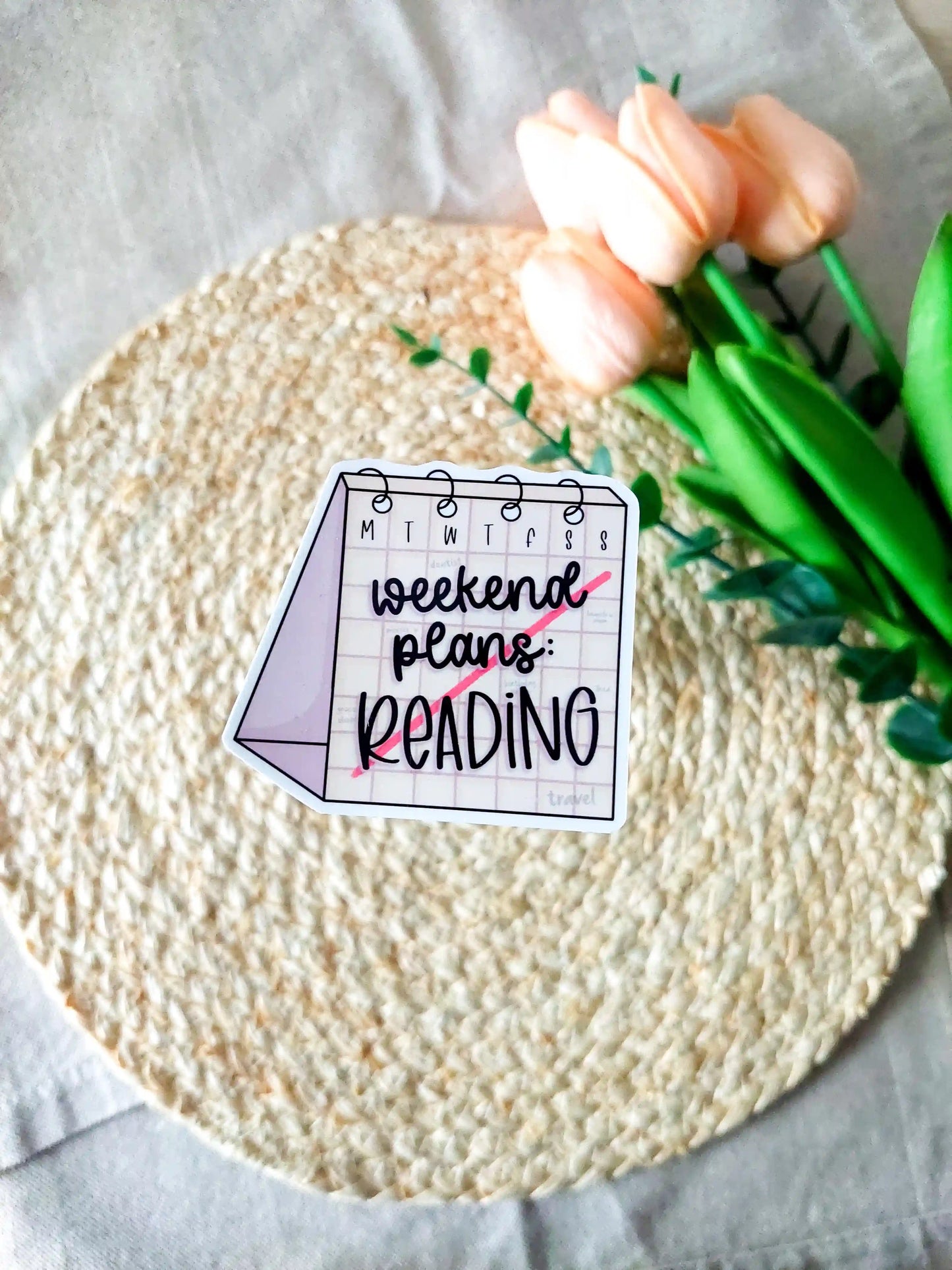 "Weekend Plans Reading" sticker on a rattan placemat, designed as a weekly calendar with black text and a line across it.
