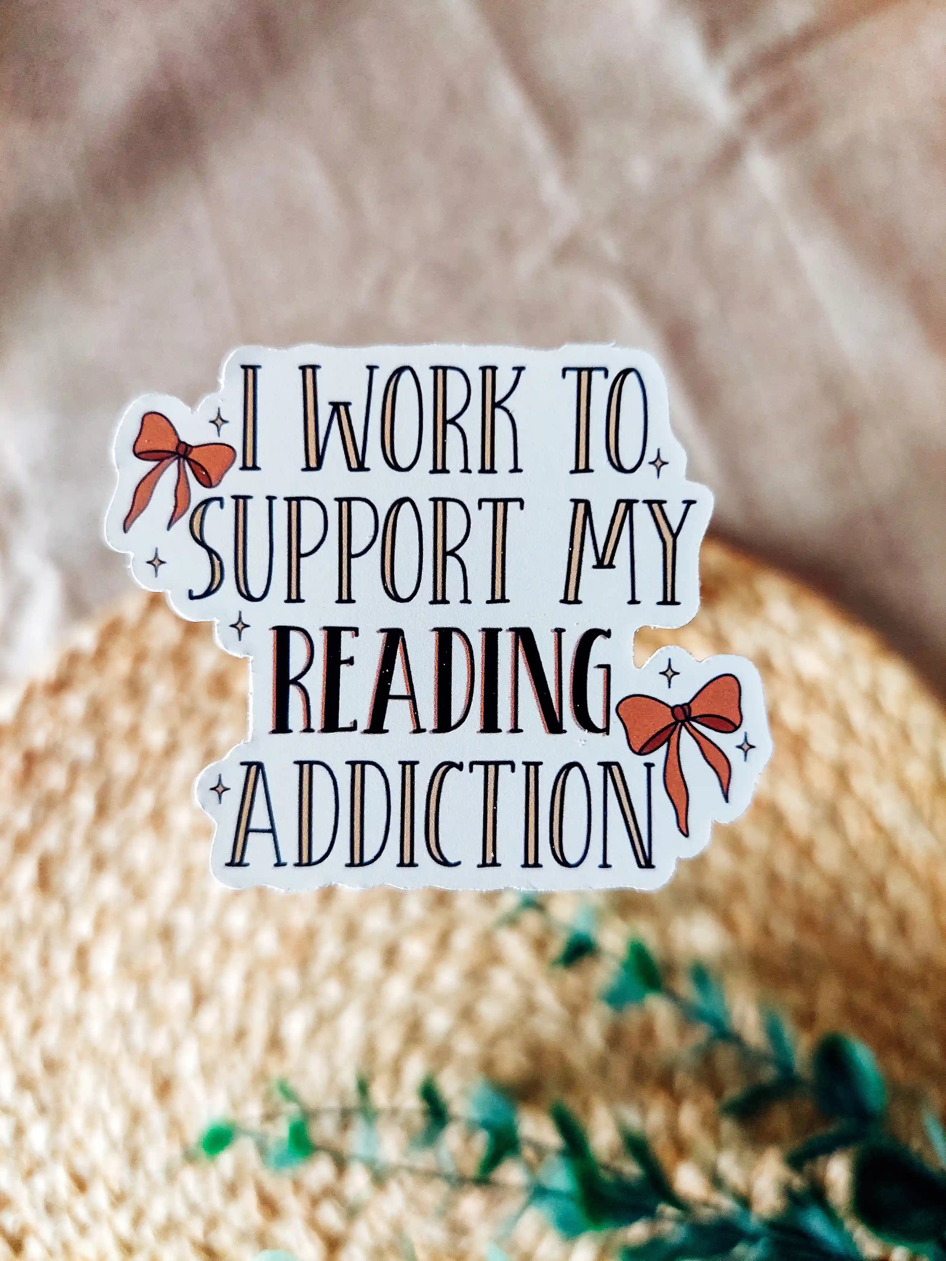 Vinyl stickers with the text "I work to support my reading addiction" with decorative orange bows, photographed on a neutral fabric background with woven mat and lavender flowers.
