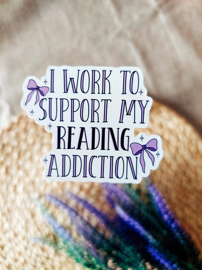 Vinyl stickers with the text "I work to support my reading addiction" with decorative purple bows, photographed on a neutral fabric background with woven mat and lavender flowers.