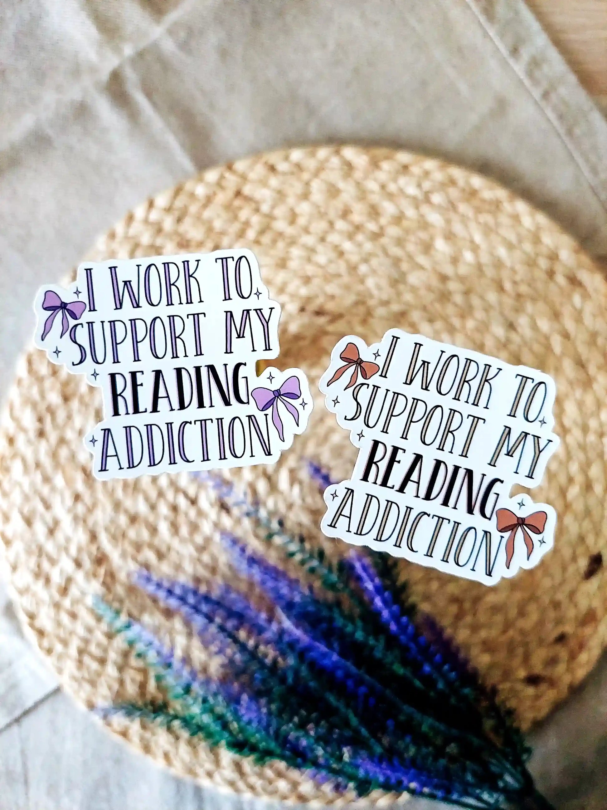 Two vinyl stickers with the text "I work to support my reading addiction" in pink and purple color variations with decorative bows, photographed on a neutral fabric background with woven mat and lavender flowers.