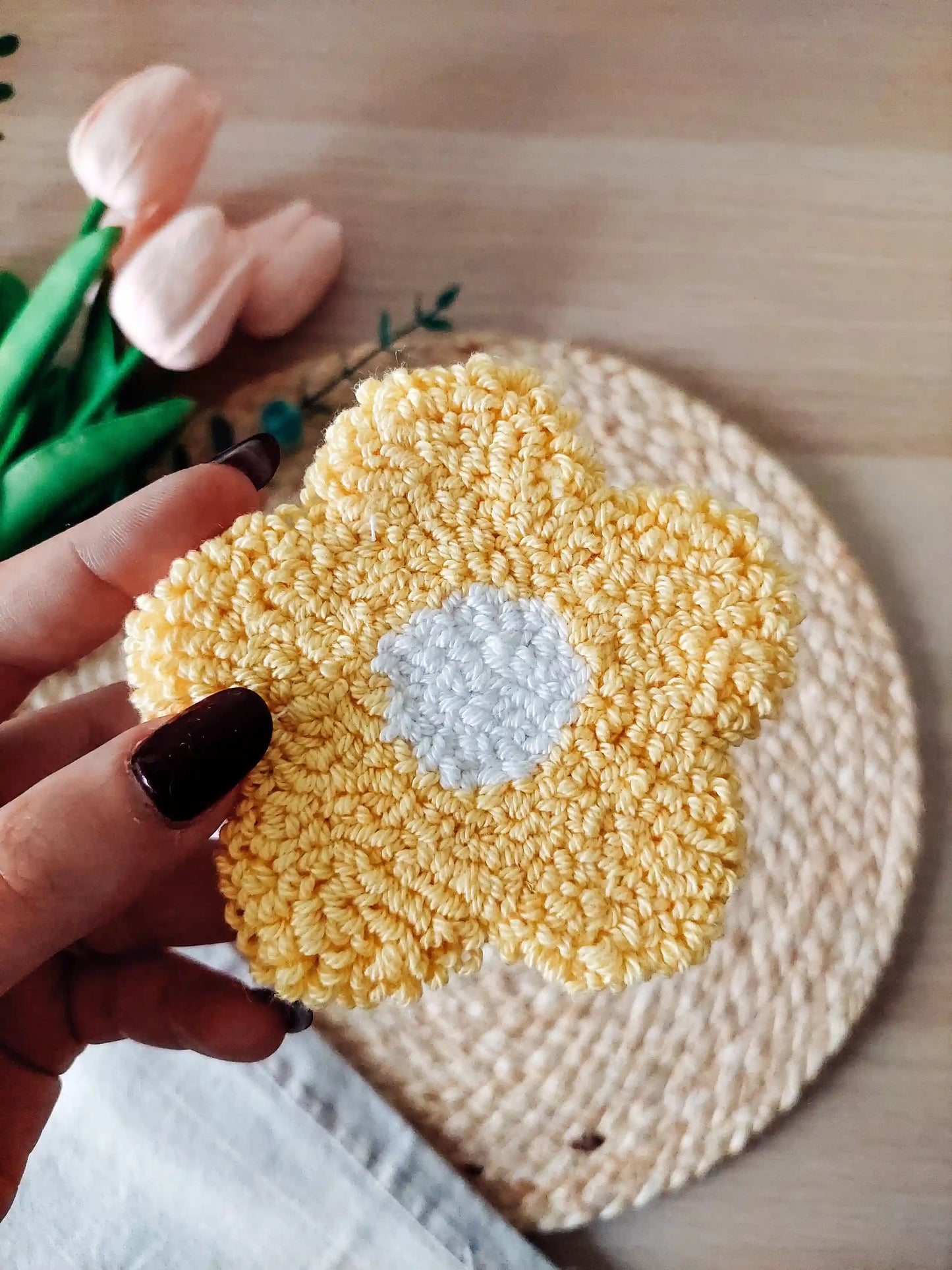Handmade punch needle coaster shaped like a yellow daisy flower with a white center, photographed on a woven mat with decorative flowers in the back.